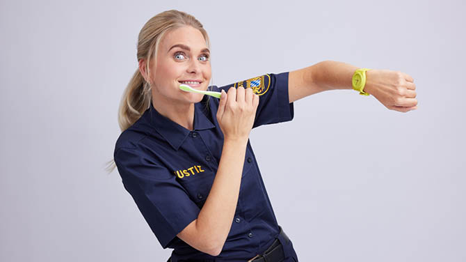 Lara K. hält ihren Arm hoch und zeigt eine Armbanduhr; gleichzeitig putzt sie ihre Zähne.